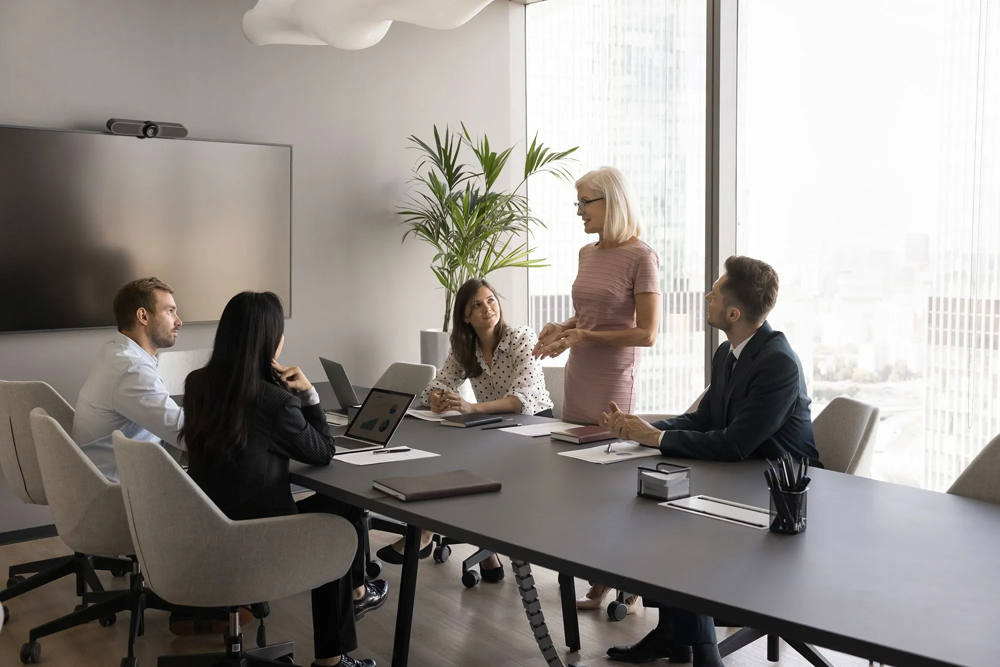 Work meeting, people sitting around a desk. Woman standing up doing a presentation.